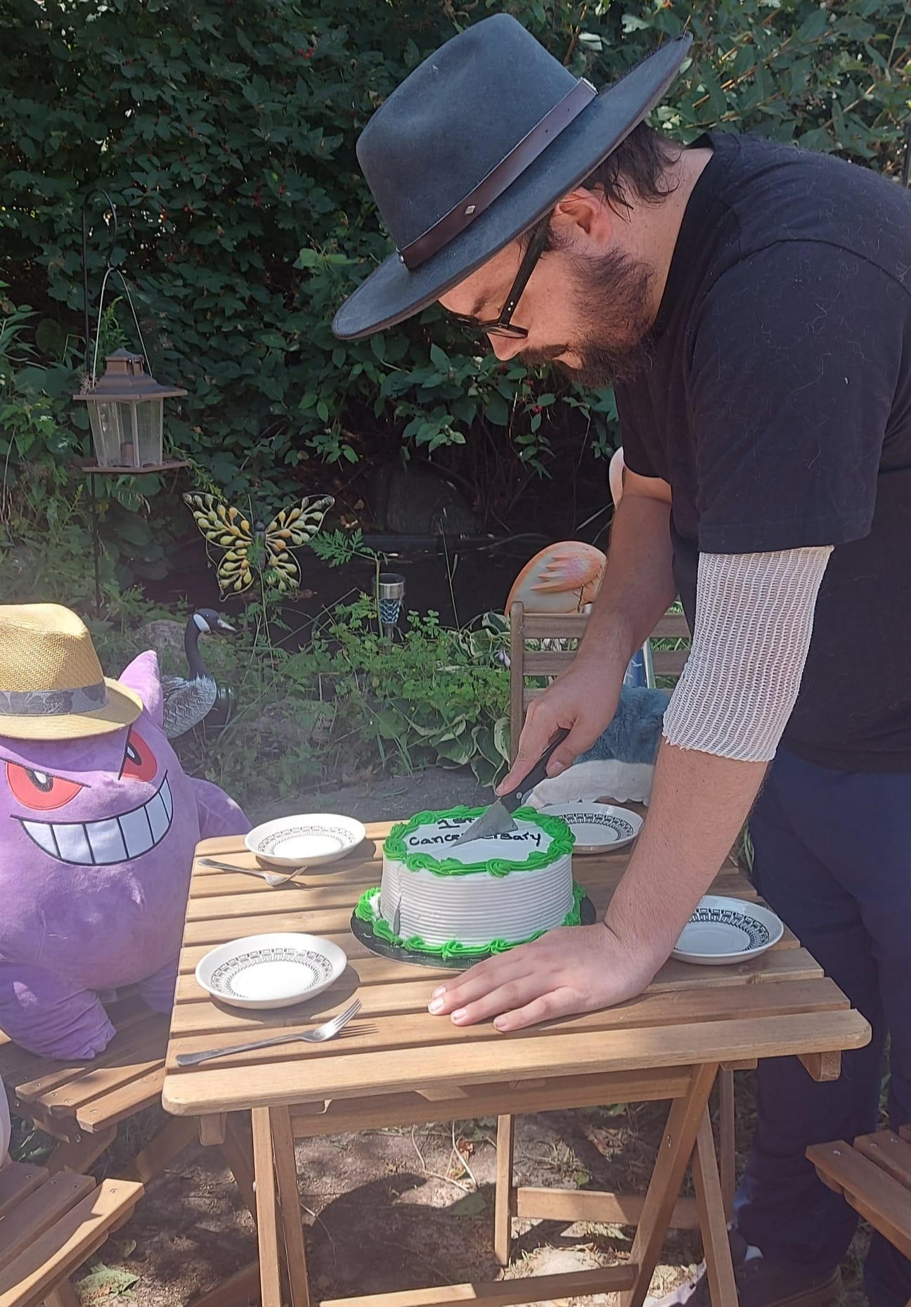 A man with facial hair and a felt sun hat has is cutting a white and green frosted cake at a stuffed animal patio party, Photo 2